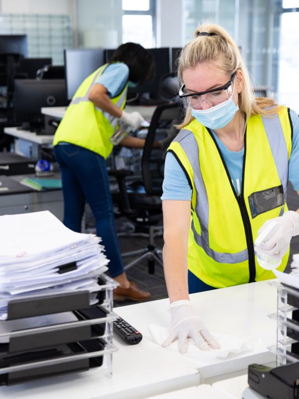 Caucasian woman and colleagues wearing hi vis vests, gloves, safety glasses and face masks sanitizing an office using disinfectant. Hygiene in workplace during Coronavirus Covid 19 pandemic.